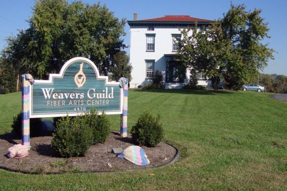 An image of the Weavers Guild house, white with a red roof and the sign in front reads Weavers Guild of Greater Cincinnati Fiber Arts Center