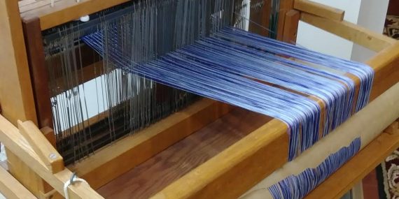 table loom with blue warp, metal heddles, levers